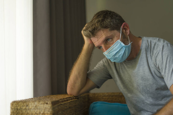covid-19 virus lockdown - sad and worried man on his 30s or 40s covered with medical mask thinking and feeling scared in quarantine following stay at home instructions to contain virus pandemic - Foto, Imagem