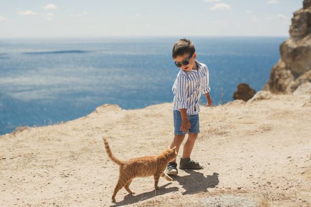 山と海を背景に赤い猫と遊ぶ幸せな少年. - 写真・画像
