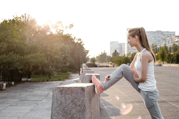 Спортивная европейская девушка в белой майке делает растяжение ног и разогрев. Молодая женщина делает упражнения на открытом воздухе - Фото, изображение