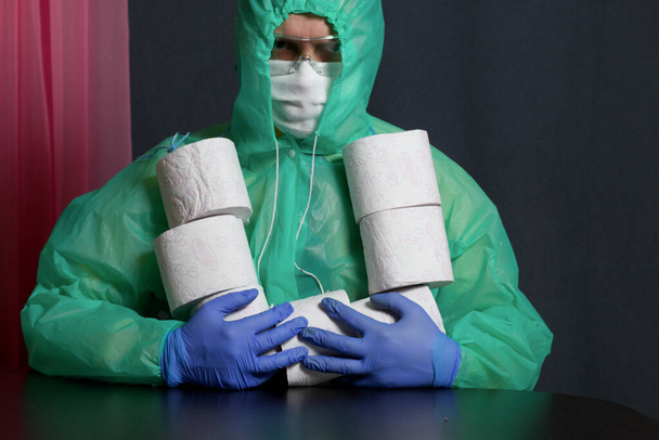 A man in a medical mask, glasses, rubber gloves and a raincoat. On the neck a bunch of rolls of toilet paper. He grabbed them with his hands. Stocks for the time of the epidemic. - Photo, Image