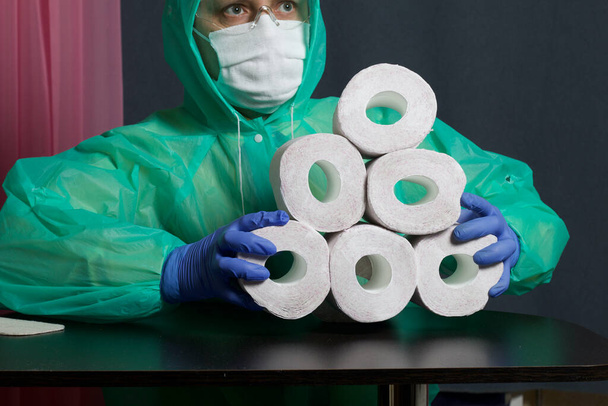 A man in a medical mask, glasses, rubber gloves and a raincoat. In the hands of rolls of toilet paper. Stocks for the time of the epidemic. - Fotoğraf, Görsel