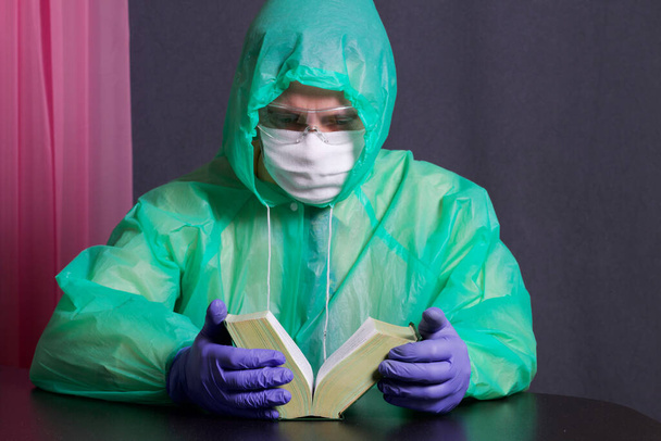 A man in a medical mask, glasses, rubber gloves and a raincoat. Front open book. Leisure during an epidemic. - Foto, Imagen