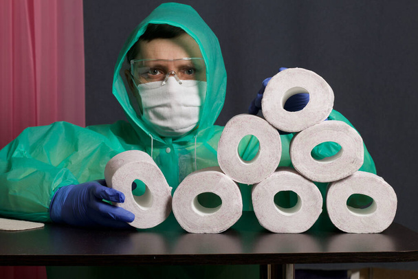 A man in a medical mask, glasses, rubber gloves and a raincoat. Folds toilet rolls on the table. Stocks for the time of the epidemic. - Photo, Image