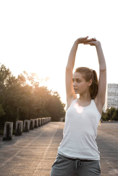 Спортивная европейская девушка в белой майке делает упражнения для рук на открытом воздухе. Девушка растягивается и разогревается - Фото, изображение