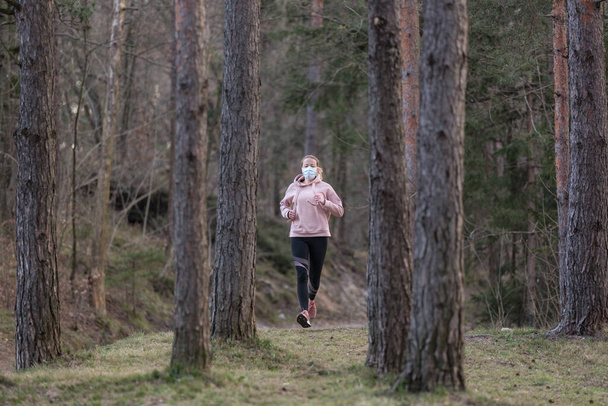 Corona virus, or Covid-19, is spreading all over the world. Portrait of caucasian sporty woman wearing a medical protection face mask while running in nature. - Foto, Imagen