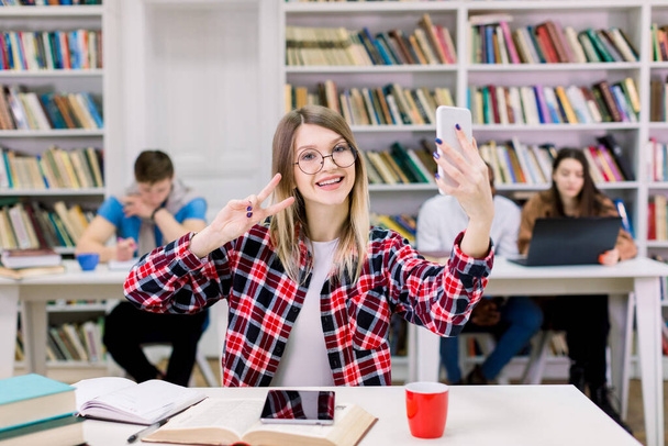 Portrét vzrušené veselé usmívající se mladé krásné blondýny v kostkované košili a brýlích dělat selfie foto a ukazující V-sign se dvěma prsty, sedí u stolu v knihovně čítárna - Fotografie, Obrázek