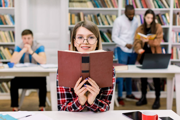 Portrait of charming smiling student girl in casual wear and eyeglasses studying at library, sitting at the table and holding open book near her face. Mixed race friends atudying on the background - Photo, Image