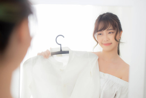 Mooie jonge aziatische vrouw glimlachen met kleren proberen op kleding passen met moderne en uitziende reflectie spiegel in de kamer, meisje kiezen kleding mode met gelukkig en vrolijk, lifestyle concept. - Foto, afbeelding