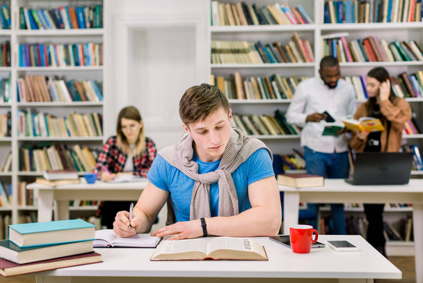 Soustředěný pohledný chlap studující a píšící poznámky z knihy, sedící u stolu v univerzitní knihovně, připravující se na zkoušky, zatímco jeho kolegové čtou knihy a diskutují na pozadí - Fotografie, Obrázek