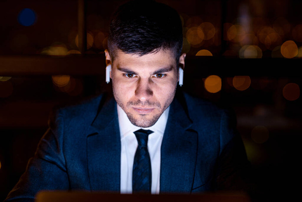 Бізнесмен у навушниках сидить на ноутбуці, працюючи пізно в приміщенні - Фото, зображення