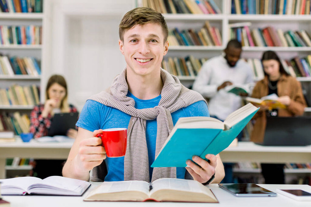 Zbliż portret radosnego, przystojnego, 25-letniego białego mężczyzny siedzącego przy stole w bibliotece, patrzącego w kamerę i pozującego z książką w jednej ręce i filiżanką herbaty w drugiej. Edukacja, koncepcja uczenia się - Zdjęcie, obraz