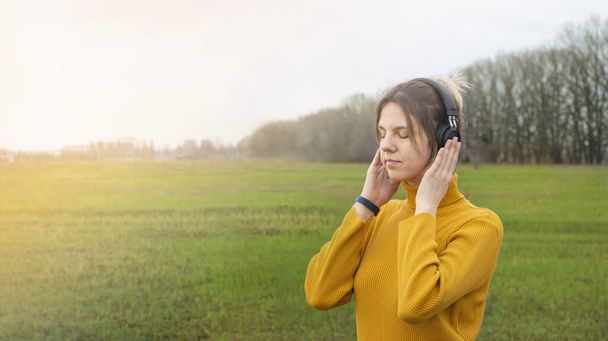 Uma jovem ouve música com fones de ouvido em um parque em um dia ensolarado de verão. Mundo moderno. Banner. Lugar para SMS. Espaço de cópia
 - Foto, Imagem
