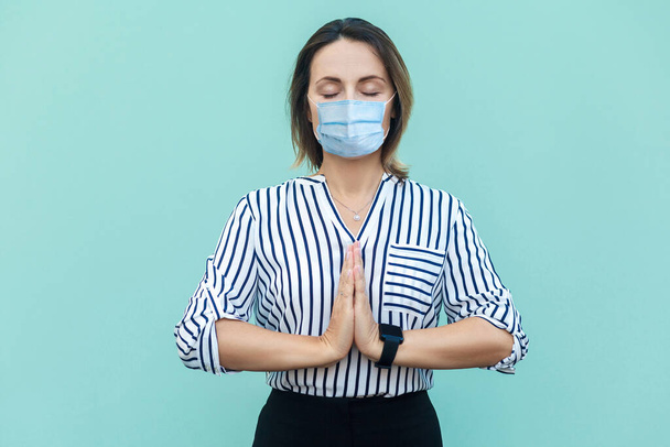 Relax and meditation. Portrait of calm middle aged woman with surgical medical mask standing and doing yoga alone. medicine and health care concept. indoor studio shot isolated on blue background. - Photo, image