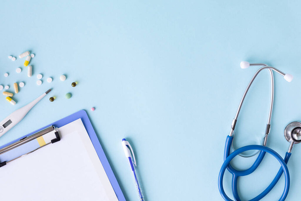 青い背景の医療概念です。聴診器、丸薬、タブレット、メモ帳、紙、ペン。医療マスク。スペースのコピー - 写真・画像