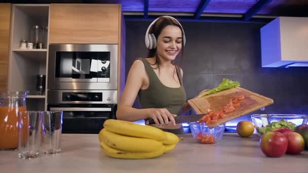 Close up of modern smiling good-looking brunette in headphones which putting into the bowl sliced tomato during preparation vegetable salad - Filmati, video