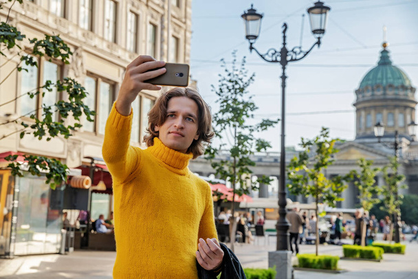 The handsome men does selfie, Look from outside, he dressed in a yellow sweater, a black raincoat or jacket is his hands, Bolshaya Konyushennaya street and Kazan Cathedral on background, sunny day - Photo, image