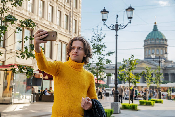The handsome men does selfie, Look from outside, he dressed in a yellow sweater, a black raincoat or jacket is his hands, Bolshaya Konyushennaya street and Kazan Cathedral on background, sunny day - Photo, Image