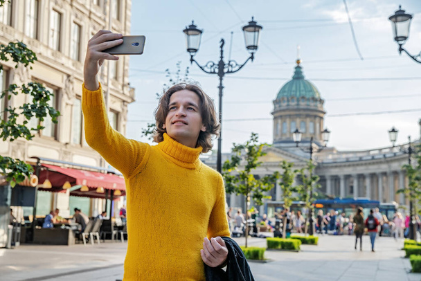 The handsome men does selfie, Look from outside, he dressed in a yellow sweater, a black raincoat or jacket is his hands, Bolshaya Konyushennaya street and Kazan Cathedral on background, sunny day - Foto, immagini