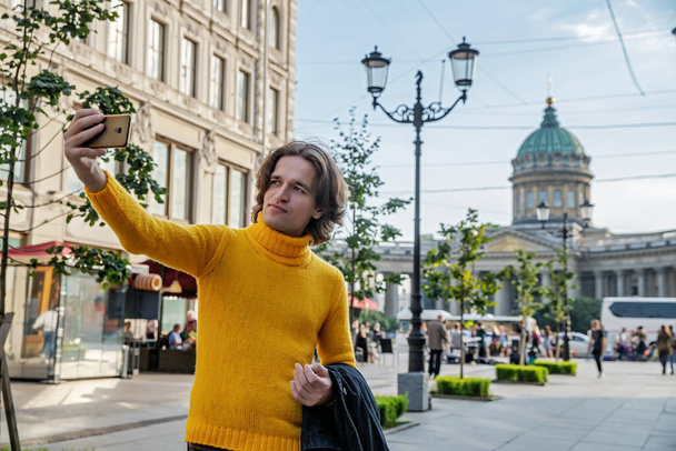 The handsome men does selfie, Look from outside, he dressed in a yellow sweater, a black raincoat or jacket is his hands, Bolshaya Konyushennaya street and Kazan Cathedral on background, sunny day - Φωτογραφία, εικόνα