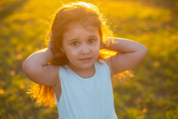cute little brunetka z długimi kręconymi włosami stoi na zielonej trawie letniego parku o zachodzie słońca i patrzy w aparat - Zdjęcie, obraz