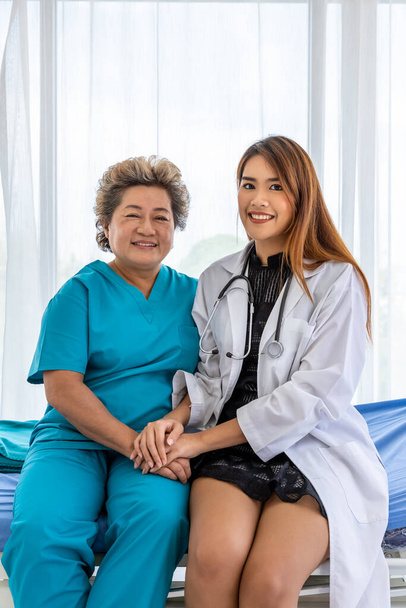 Asian medical doctor holding senior elderly woman female patient hand sitting on bed to comfort and motivate, looking at camera - Photo, Image