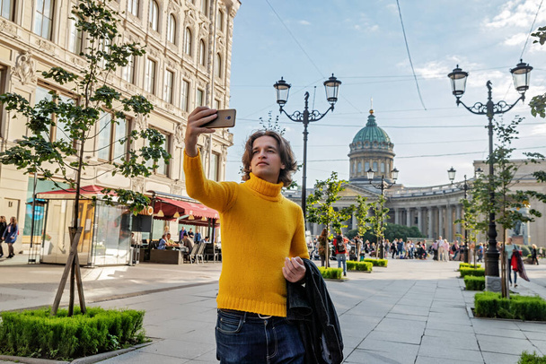 De knappe mannen doet selfie, Kijk van buiten, hij gekleed in een gele trui, een zwarte regenjas of jas is zijn handen, Bolsjaja Konyushennaya straat en Kazan kathedraal op de achtergrond, zonnige dag - Foto, afbeelding