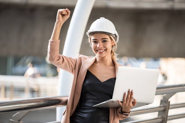 Happy 20s American engineer woman with helmet and coat working on laptop raise hand at urban city. Beautiful female worker celebrate construction project or win bid auction from internet. - Photo, Image