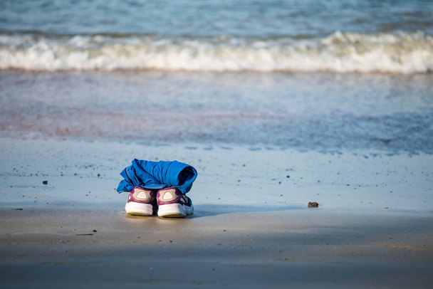 Calzado y t-shrirt azul en la arena en la playa por la mañana. Zapatos y camisa junto al mar con movimiento borroso ola oceánica. Concepto de vacaciones de verano
. - Foto, Imagen