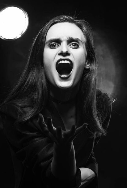 Beautiful braless girl with red lips, wearing a black blazer, screams, stretching forward her hands on a dark background, posing next to a light lamp. Advertising, trendy, monochrome design - Foto, afbeelding