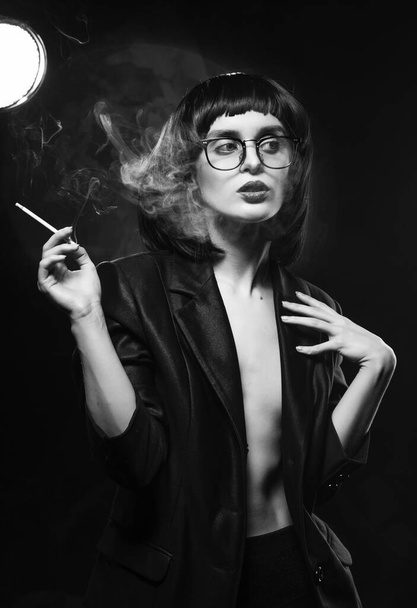 Beautiful braless slim girl with red lips, wearing a unbuttoned black blazer, glasses and brunette wig sensually smokes un a dark background next to the light lamp. Artistic monochrome noir design - Foto, imagen