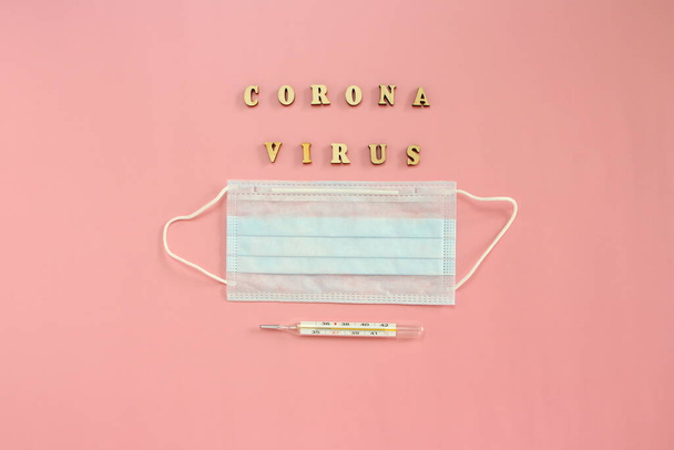 Medical disposable mask, thermometer and wooden letters forming the word Coronovirus on a pink background - Foto, imagen