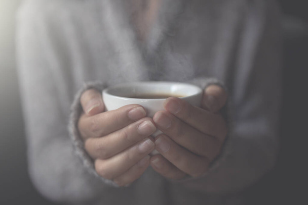 Großaufnahme weiblicher Hände mit einer Tasse heißen Kaffees. am Morgen sanftes Sonnenlicht. Schöne Mädchen im grauen Pullover. - Foto, Bild