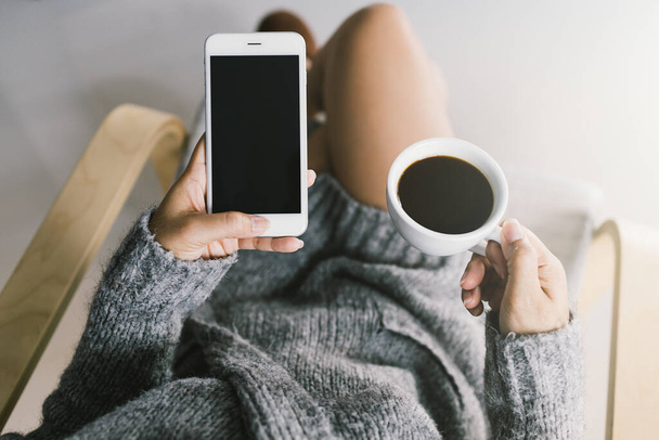 Mockup immagine delle mani della donna che tiene il telefono cellulare schermo nero con schermo bianco e tazza di caffè in mano
. - Foto, immagini