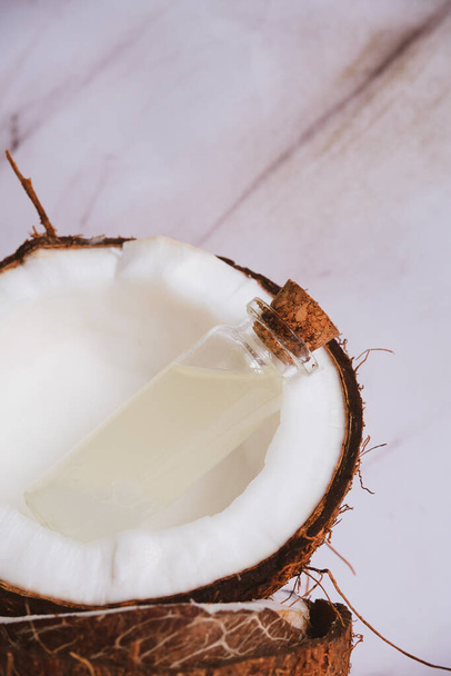 close up selective focus glass bottle with coconut oil on a blurred background of cracked coconuts - Фото, изображение