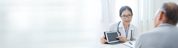 Beautiful Asian Female Doctor talking some information about virus to elderly patient with digital tablet. Physician or Practitioner wear glasses giving advice to senior man in medical room of hospital. Banner, Copy space, Panoramic. - Foto, Bild
