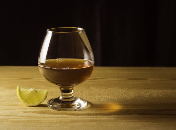 Γυάλινο ποτήρι με κονιάκ σε ξύλινο τραπέζι, φέτα λεμονιού, σκούρο φόντο. Ελεύθερος χώρος για κείμενο στο πλάι. - Φωτογραφία, εικόνα