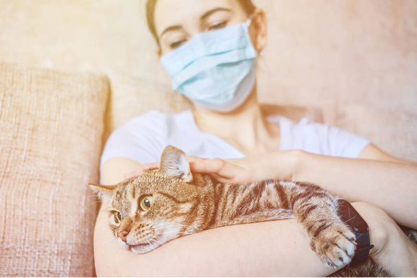 A girl in a medical mask is in quarantine at home petting a cat in her arms. Close up. - Photo, Image