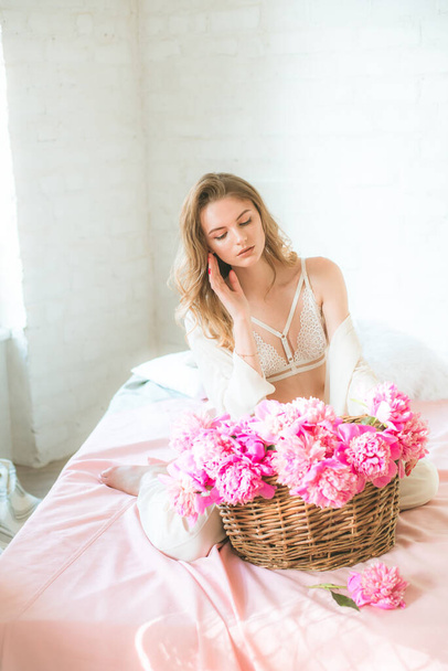 Cute tender young girl with blond hair in white clothes and lace underwear with a basket of pink peonies on the bed in the studio. Spring and flowers. Beauty and Fashion - Foto, Bild