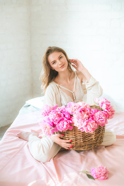Cute tender young girl with blond hair in white clothes and lace underwear with a basket of pink peonies on the bed in the studio. Spring and flowers. Beauty and Fashion - Φωτογραφία, εικόνα