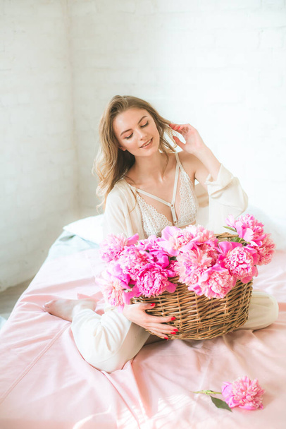 Cute tender young girl with blond hair in white clothes and lace underwear with a basket of pink peonies on the bed in the studio. Spring and flowers. Beauty and Fashion - Фото, зображення
