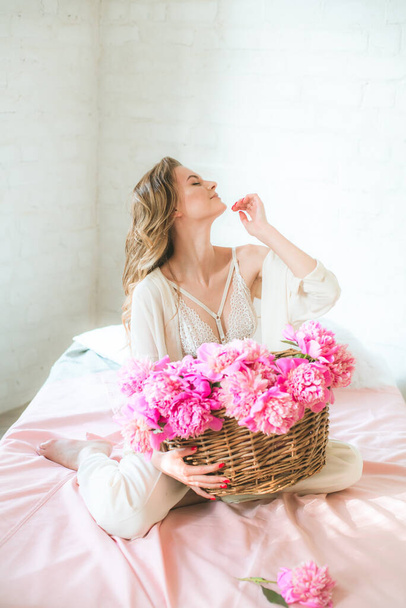 Cute tender young girl with blond hair in white clothes and lace underwear with a basket of pink peonies on the bed in the studio. Spring and flowers. Beauty and Fashion - Foto, Imagen