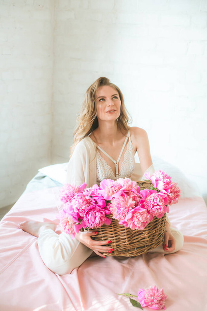 Cute tender young girl with blond hair in white clothes and lace underwear with a basket of pink peonies on the bed in the studio. Spring and flowers. Beauty and Fashion - 写真・画像