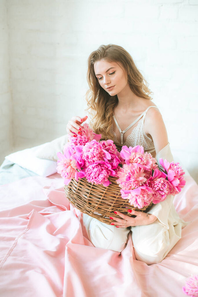 Cute tender young girl with blond hair in white clothes and lace underwear with a basket of pink peonies on the bed in the studio. Spring and flowers. Beauty and Fashion - Photo, Image