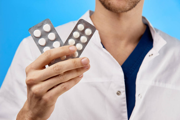 De dokter houdt in zijn hand een blaren met pillen, toont medicijnen in de camera close-up geïsoleerd op een blauwe achtergrond - Foto, afbeelding