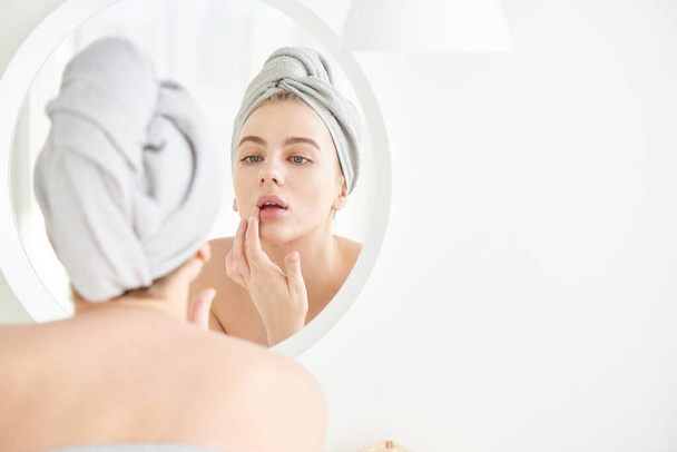 Portrait of  young girl with  towel on head in white bathroom looks and touches her face in the mirror and enjoys youth and hydration. Natural beauty, home care for problem skin - Fotó, kép