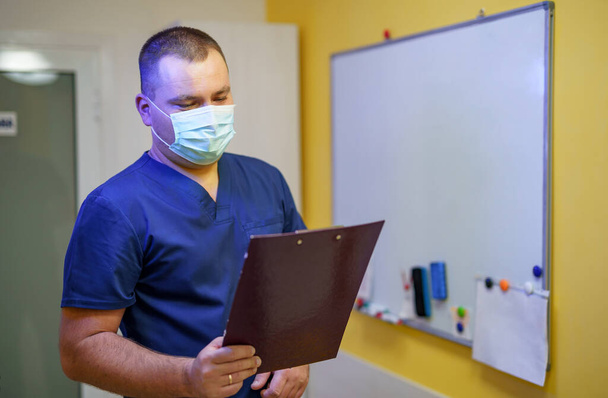 Серьезный хирург в маске, стоящий в офисе с папкой
. - Фото, изображение