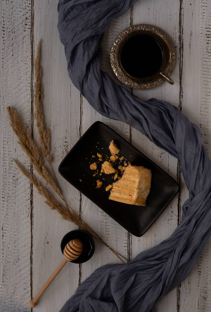 Sweet homemade layered honey cake on a black plate, white table, with honey and coffee. - Photo, Image