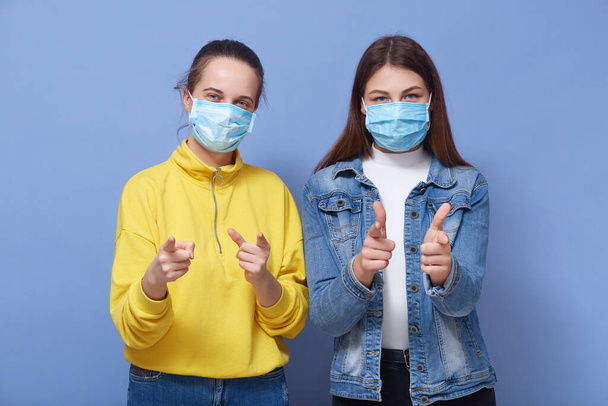 Indoor studio picture of two charismatic good looking females wearing protection masks, making gestures, showing direction with forefingers, standing isolated over blue background. Covid 19 concept. - 写真・画像