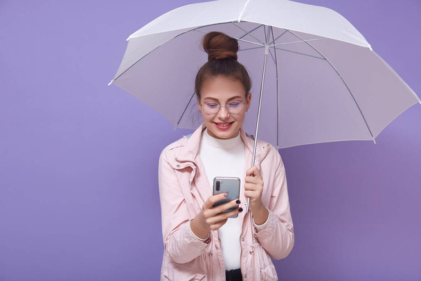 Indoor picture of smiling positive model with bun holding umbrella and smartphone, having peaceful facial expression, using her device actively, spending time alone. Youth and technology concept. - Photo, Image