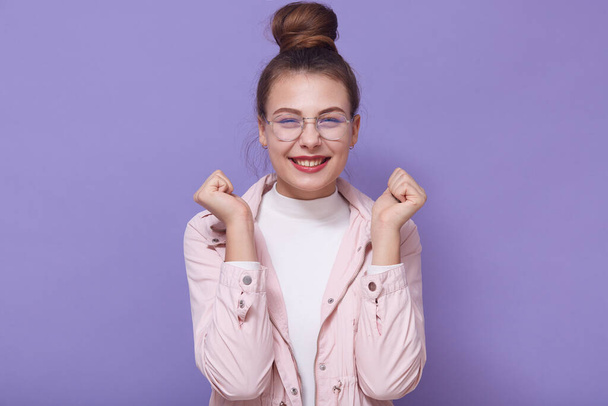 Studio image of happy funny joyful young lady keeping hands in fists, having success, being in good mood, wearing accessories, having cute smile, enjoying free time alone. Spring time concept. - Fotó, kép